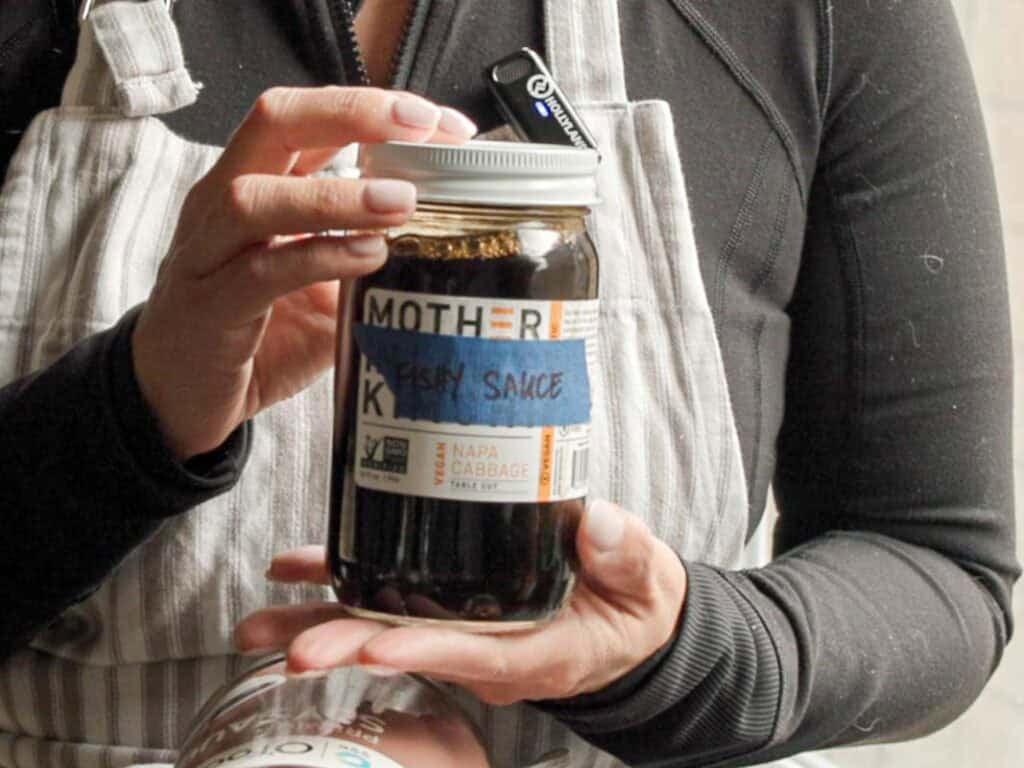 Person holding a jar labeled Fishy Sauce with “Mother” and “Napa Cabbage” on a sticker. They are wearing a black top and striped apron. Another jar is partially visible at the bottom.